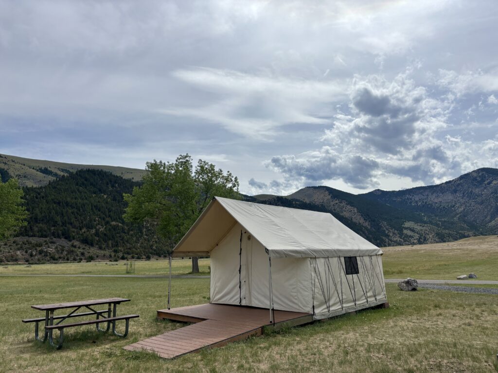 Canvas wall tent accommodation at Lewis and Clark Caverns State Park campground in Southwest Montana, set on a wooden platform with a picnic table in the foreground and the Jefferson River Valley mountains rising behind it under a partly cloudy sky.