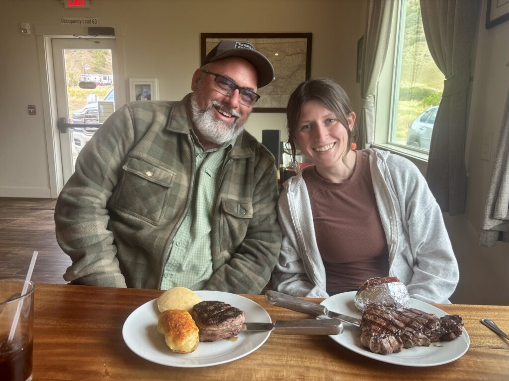 The author and her father smile at a wooden table inside LaHood Park Steakhouse in Cardwell, Montana, with two locally sourced Montana beef dinners in front of them, a New York Strip served with a loaded baked potato and a Tenderloin served with a dinner roll and twice-baked potato.