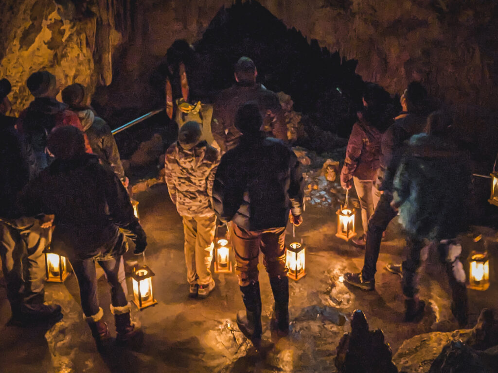 A group of visitors stands in a dark cave chamber holding glowing candle lanterns during the Candlelight Tour at Lewis and Clark Caverns State Park in December, the warm lantern light illuminating wet cave floors and towering stalactite formations overhead.
