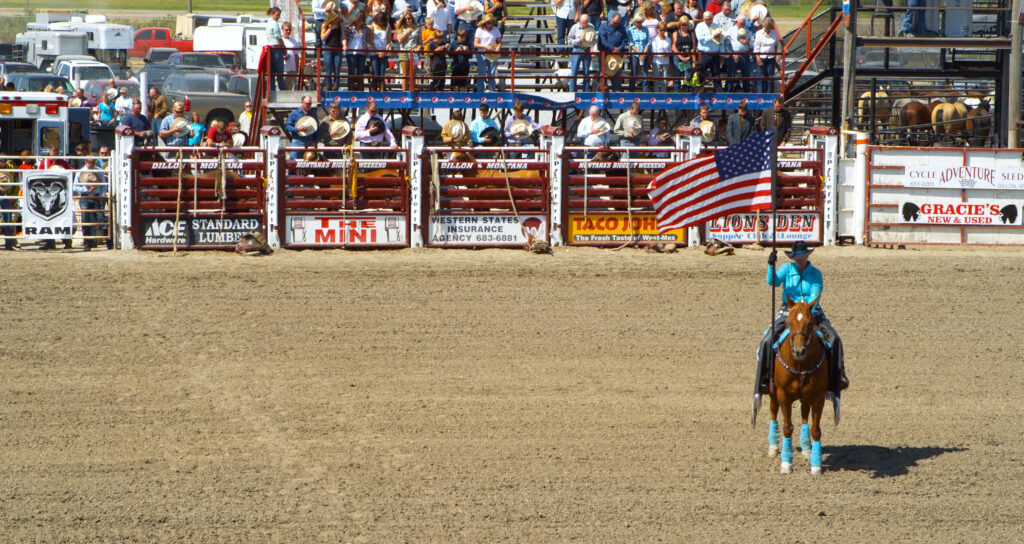 Flag bearer on horseback leads the grand entry at the Dillon Jaycees PRCA Rodeo, part of Montana's Biggest Weekend in Dillon, Montana. Spectators pack the grandstand behind sponsor-lined rodeo chutes, capturing the Labor Day weekend rodeo tradition in Southwest Montana. Photo by Tempest Technologies LLC.