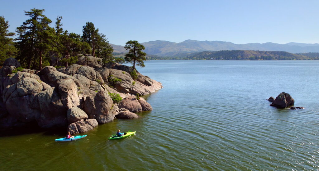 Two kayakers paddle along a rocky, pine-covered shoreline on Canyon Ferry Lake near Helena, Montana, with calm water and distant mountains in the background. The rugged granite outcrops are a popular spot for exploring by kayak or paddleboard. Photo by Mike Dreesman, Tempest Technologies.