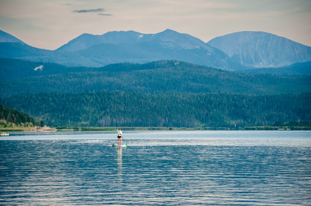 A paddleboarder glides across the calm, glassy water of Georgetown Lake, Montana, with dense pine forest and the layered peaks of the Pintler Mountains rising in the background. The stillness of the lake makes it a favorite spot for paddleboarding at dusk. Photo by Lisa Edwards.