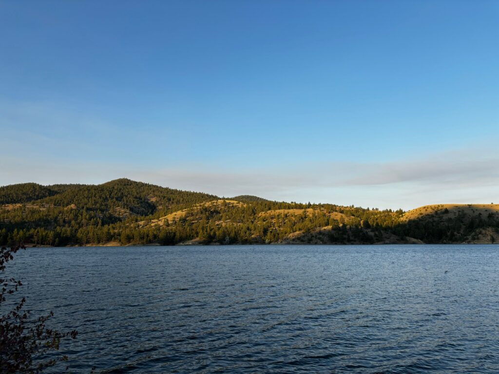 Evening light hits pine-covered hills along the shoreline of Hauser Lake, Montana, as seen from Black Sandy State Park, with calm blue water stretching toward rolling terrain in the distance. This quieter reservoir north of Helena offers easy access for boating, fishing, and swimming. Photo by Alyssa Starr, Tempest Technologies.