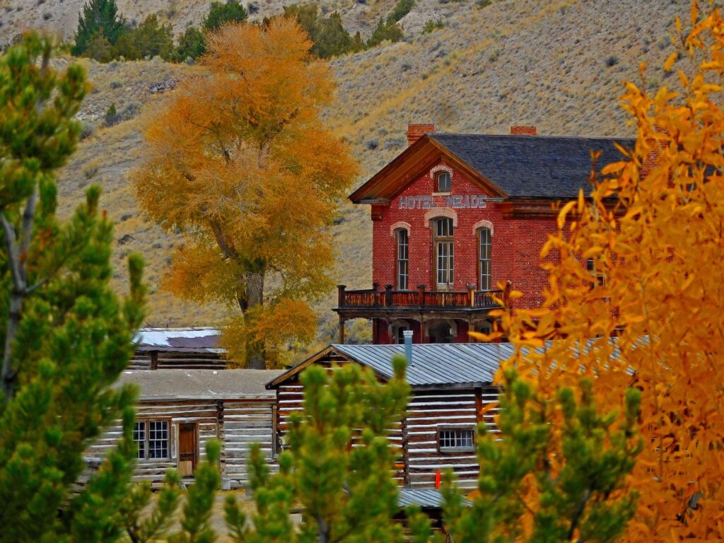 The red brick Hotel Meade, with its name lettered across the front and a wooden balcony above the entrance, rises behind weathered log cabins at Bannack State Park in Montana. A large cottonwood turns golden orange beside the hotel, bright orange leaves frame the right side of the scene, and green pine branches fill the foreground. A sagebrush hillside dotted with junipers rises behind the ghost town, a fall foliage scene in Southwest Montana. Photo by Roger Kasak, Bannack State Park.
