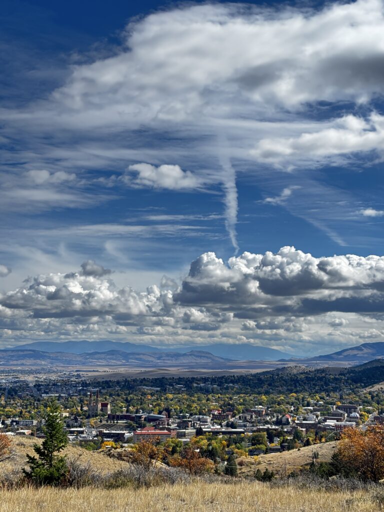 View of downtown Helena, Montana, from Mount Helena City Park in early October, with fall colors in yellow, gold, and green across the city's tree canopy. The twin spires of the Cathedral of St. Helena rise above brick buildings on the left, while orange and rust-colored shrubs and a young conifer stand in golden grass in the foreground. Beyond town, the Helena Valley stretches toward distant mountains under a deep blue sky full of white cumulus and wispy clouds, a classic fall foliage view in Southwest Montana. Photo by Alyssa Starr, Tempest Technologies LLC.