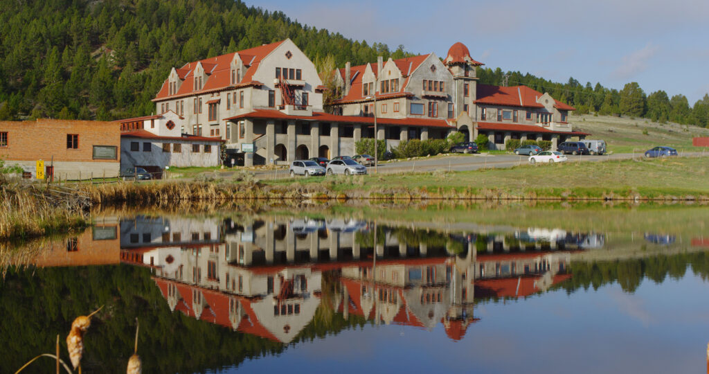 Historic Boulder Hot Springs near Boulder, Montana, a sprawling Spanish Mission-style hotel with gray stucco walls, red tile roofs, stepped gables, an arched colonnade, and a domed tower set against a pine-covered hillside. The full building reflects in a calm pond in the foreground, framed by dry cattails and green spring grass under a soft blue sky. Photo by Mike Dreesman, Tempest Technologies LLC