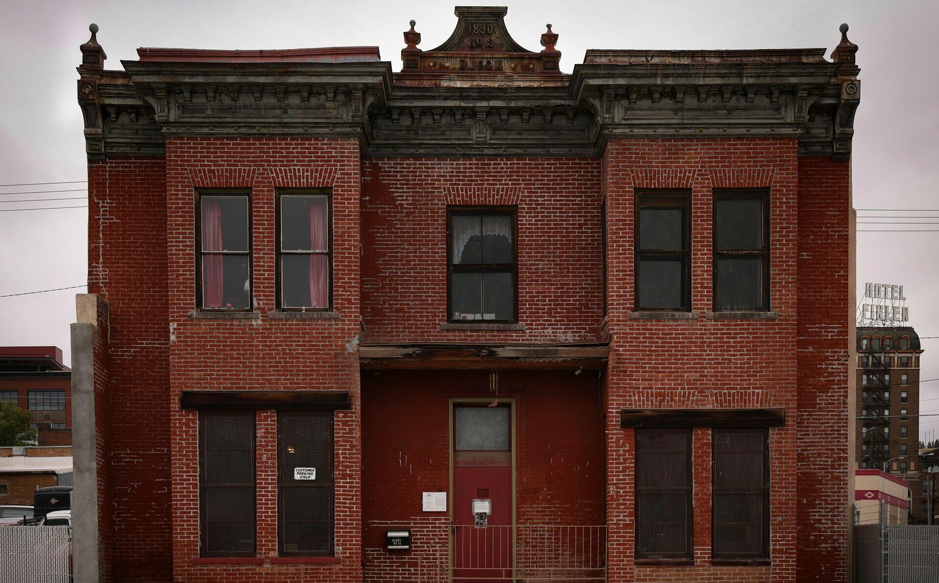 Front facade of the historic Dumas Brothel on East Mercury Street in Uptown Butte, Montana, a two-story red brick building with an ornate pressed-metal cornice, decorative finials, and a weathered pediment marked 1890 No. 2 Block. Red curtains hang in the upstairs windows above a red front door with an iron railing, while the rooftop Hotel Finlen sign rises in the background under an overcast sky. Once a brothel on the edge of Butte’s Venus Alley red-light district, the Dumas is said to be one of the most haunted buildings in Southwest Montana. Photo by Sarah Bolt, Tempest Technologies