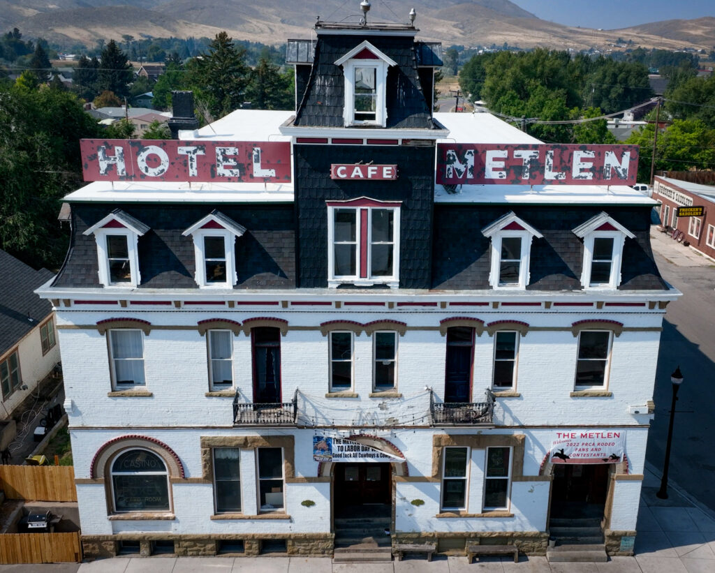 Aerial view of the historic Hotel Metlen in Dillon, Montana, a three-story Second Empire-style landmark with white-painted brick, a dark-shingled mansard roof, white dormer windows, and a central tower with a small CAFE sign. Weathered red-and-white HOTEL METLEN signs stretch across the rooftop, two wrought-iron balconies sit above arched entryways hung with rodeo welcome banners, and golden foothills rise beyond the trees in the background. Photo by Sarah Bolt, Tempest Technologies LLC