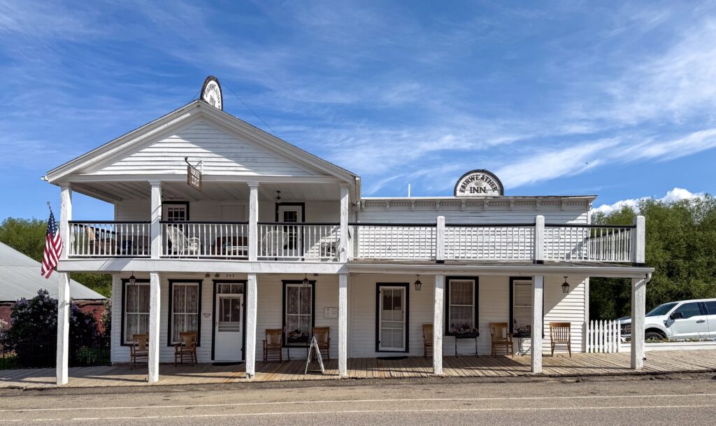 Historic Fairweather Inn on Wallace Street in Virginia City, Montana, a white two-story wooden hotel with a front-gabled entrance, an upper balcony lined with white wicker chairs, and a covered wooden boardwalk with slat-back chairs facing the street. Arched Fairweather Inn signs top the roofline, an American flag hangs from a porch post, and purple lilacs bloom beside the building under a bright blue sky with wispy clouds. Photo by Sarah Bolt, Tempest Technologies LLC