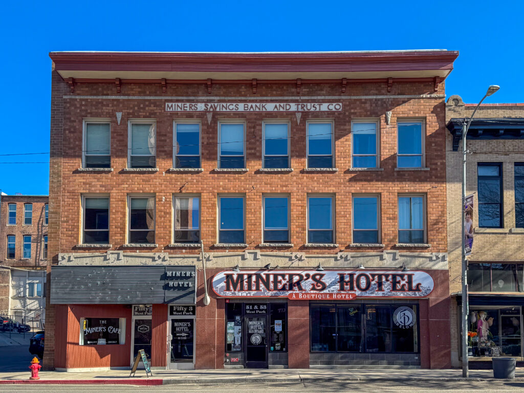 Historic Miner’s Hotel on West Park Street in Uptown Butte, Montana, a three-story red brick building with a bracketed cornice and the original MINERS SAVINGS BANK AND TRUST CO. lettering carved above two rows of tall windows. At street level, a large Miner’s Hotel A Boutique Hotel sign stretches over the main entrance beside The Miner’s Cafe storefront, with a red fire hydrant on the sidewalk under a clear blue sky. Photo by Sarah Bolt, Tempest Technologies LLC